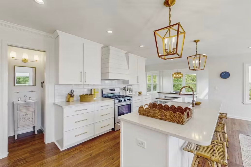 White kitchen with island, cabinets, and pendant lights. Countertops, stove, and a view into a hallway with a bathroom. White kitchen with island, cabinets, and pendant lights. Countertops, stove, and a view into a hallway with a bathroom.