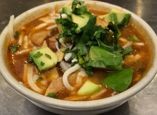 A bowl of soup with noodles , avocado , and cilantro on a table.