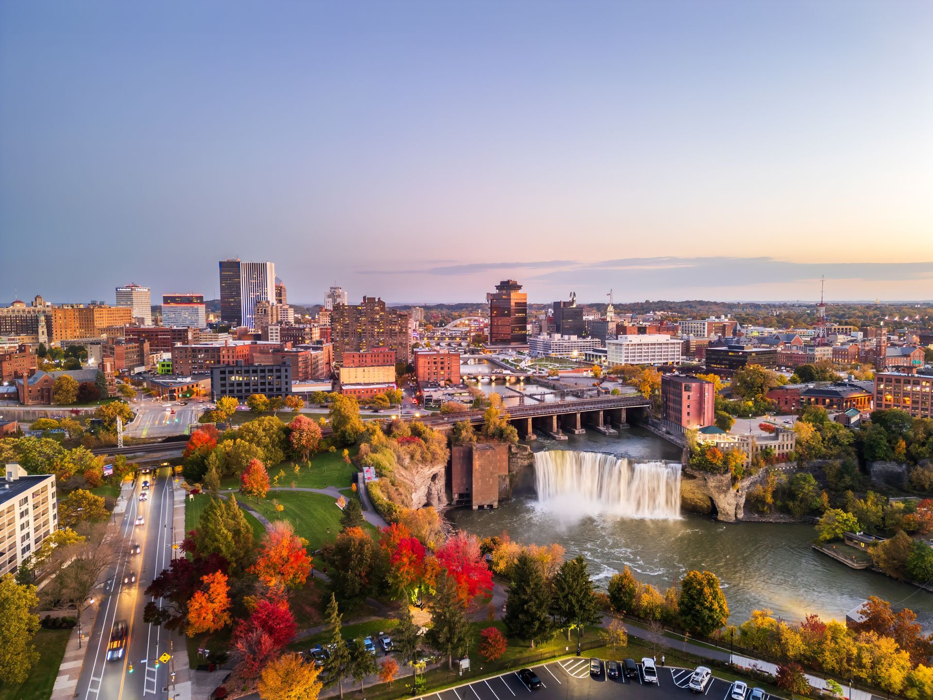 Aerial view of Rochester, NY, with Genesee River waterfall in foreground, autumn foliage, and city skyline.