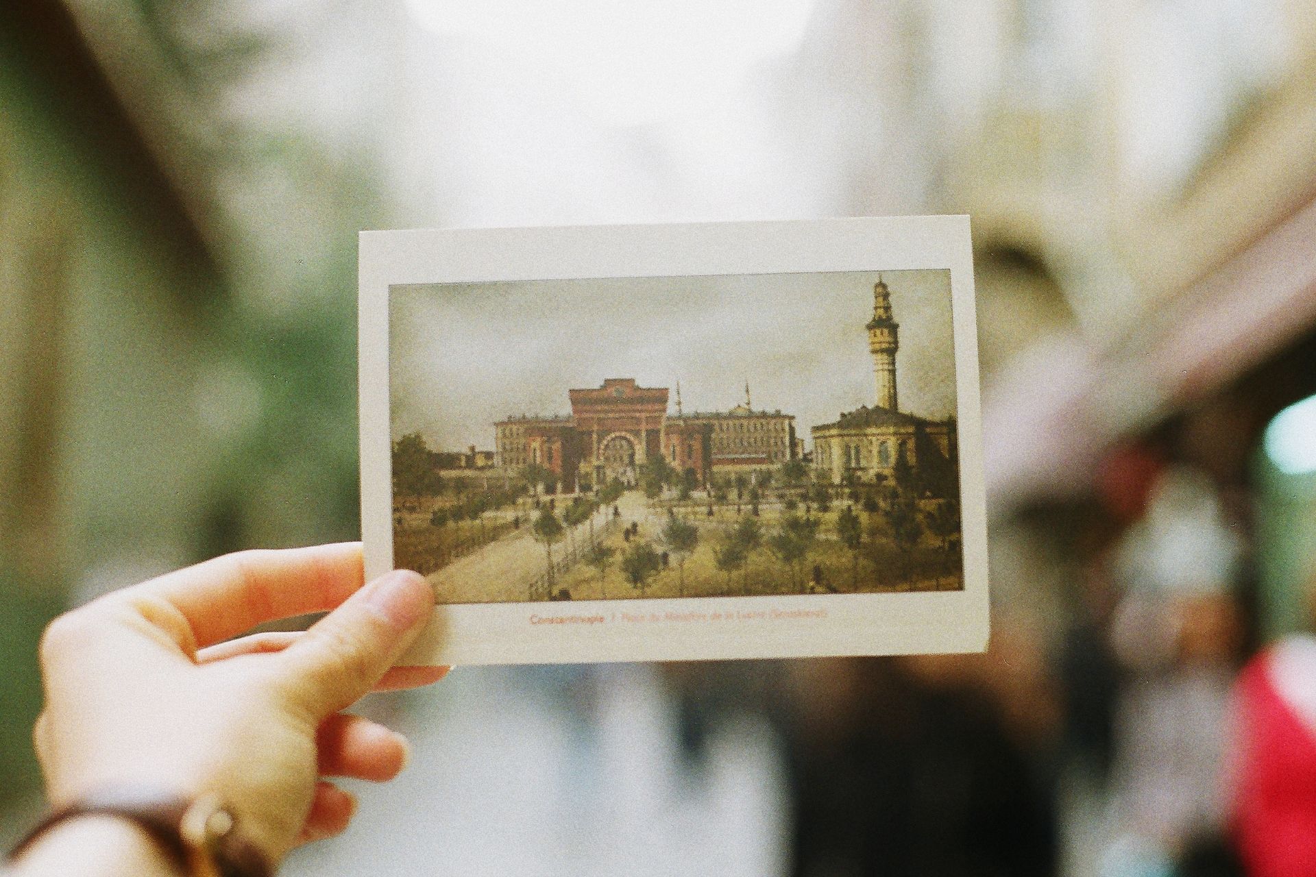 Hand holding postcard of a historic building with a tower, blurred street scene in background.