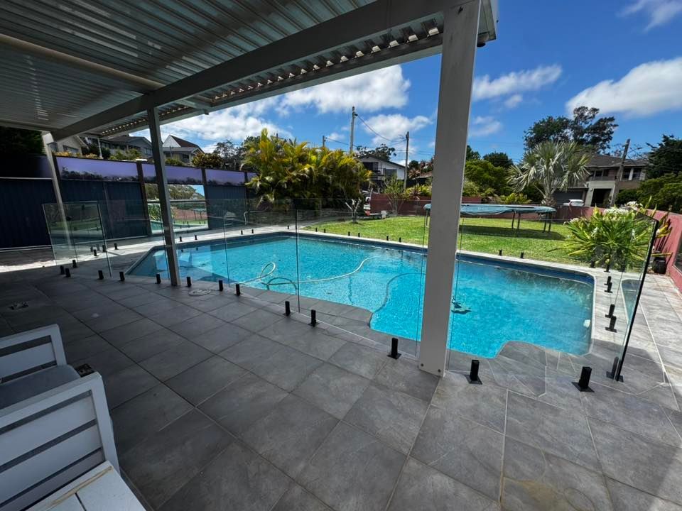 Covered outdoor pool with clear blue water, glass railing, and patio seating under a roof — ClearView Glass Pool Fencing In Roselands, NSW