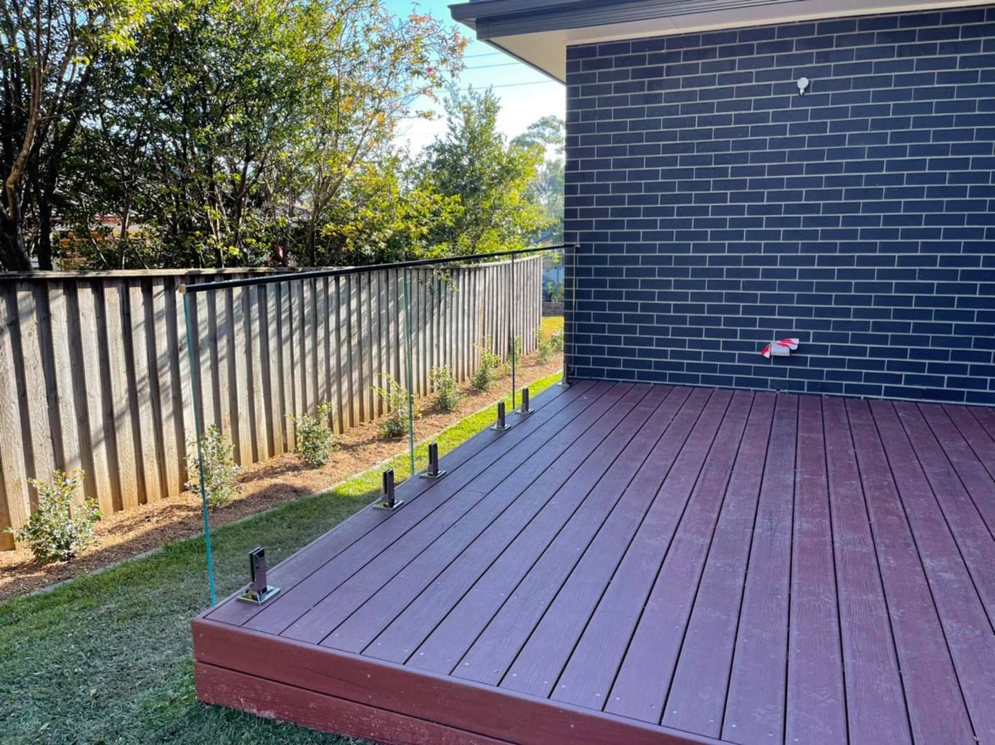 Backyard deck with red flooring beside a dark brick wall and a metal fence, with trees beyond — ClearView Glass Pool Fencing In Roselands, NSW