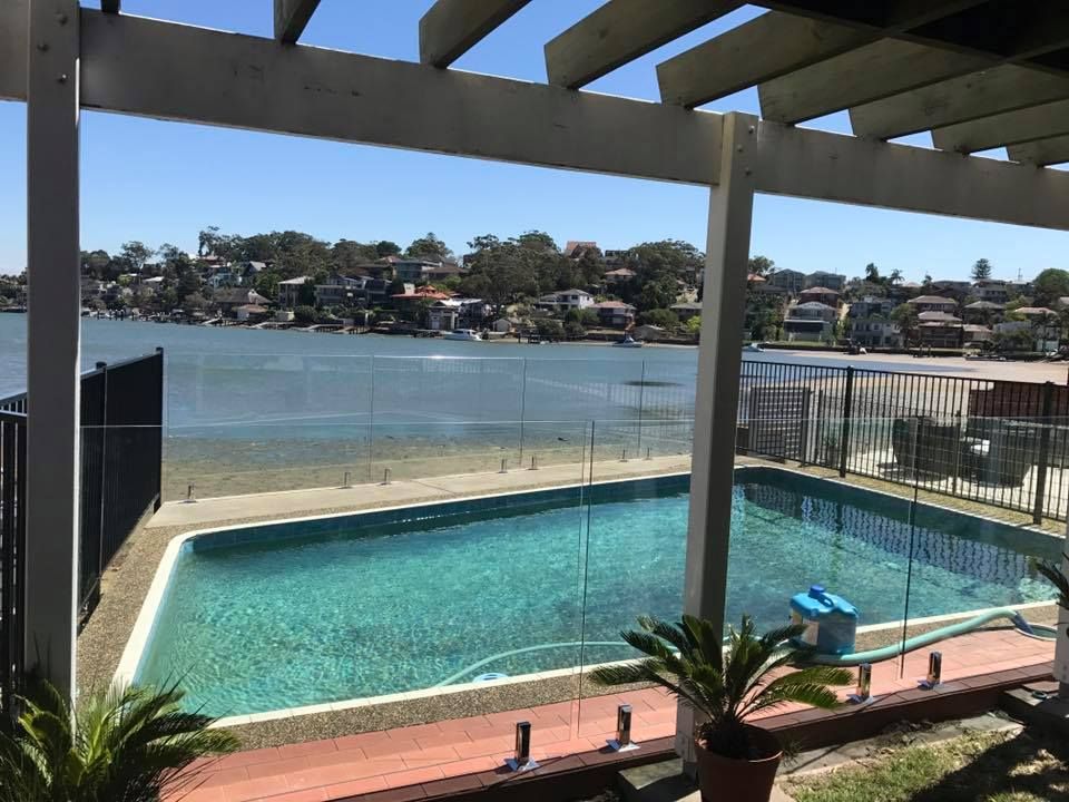 Backyard pool with pergola, overlooking a calm bay and hillside homes — ClearView Glass Pool Fencing In Roselands, NSW