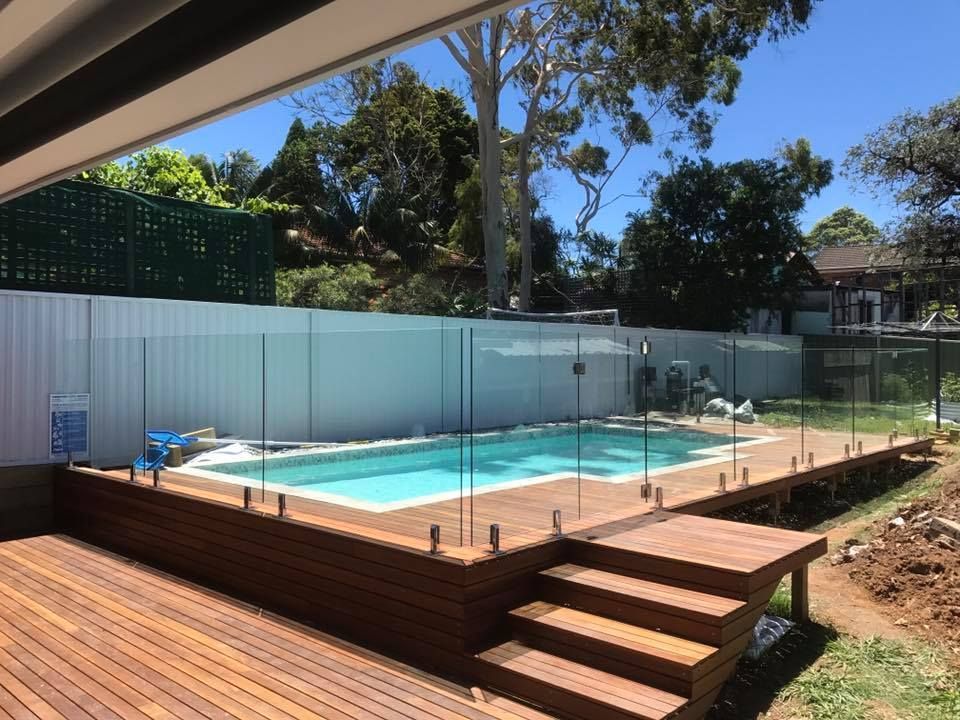 Backyard swimming pool with glass fence, wooden deck, and steps under a blue sky — ClearView Glass Pool Fencing In Roselands, NSW