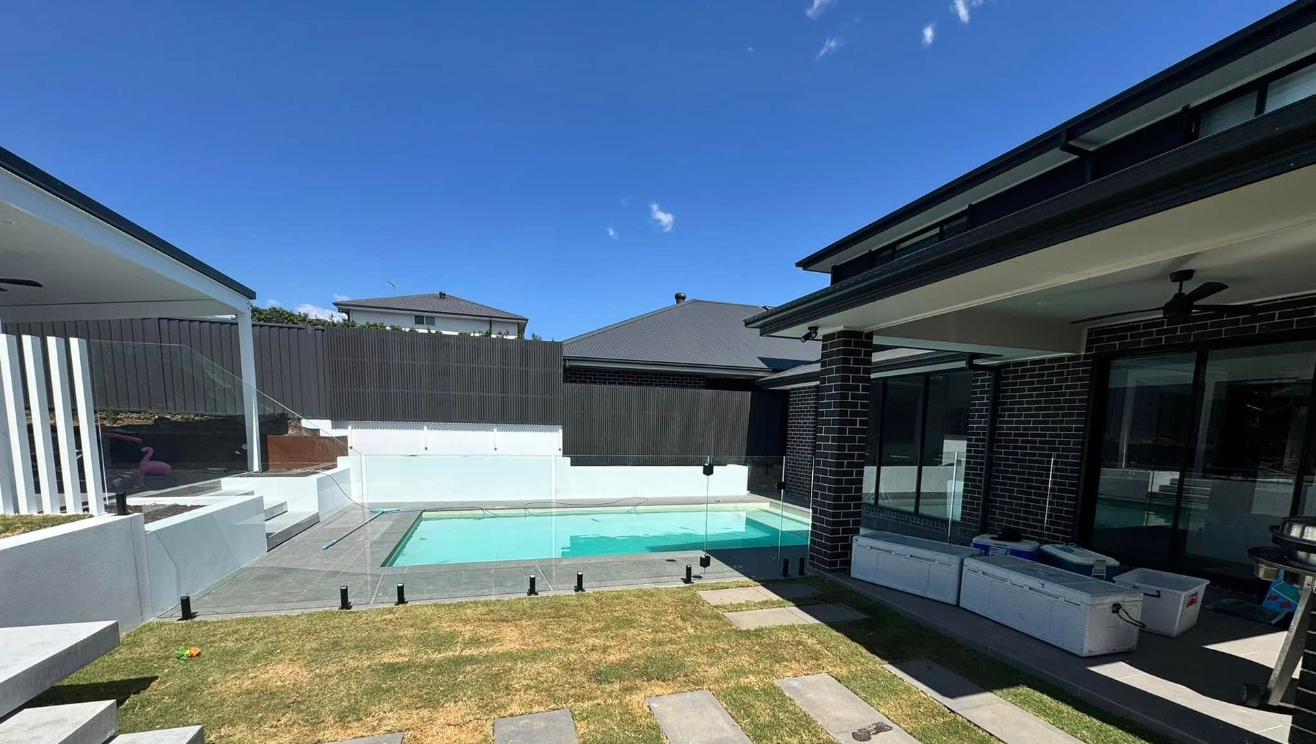 Backyard patio with a turquoise swimming pool, glass fence, and modern houses under a clear blue sky — ClearView Glass Pool Fencing In Roselands, NSW