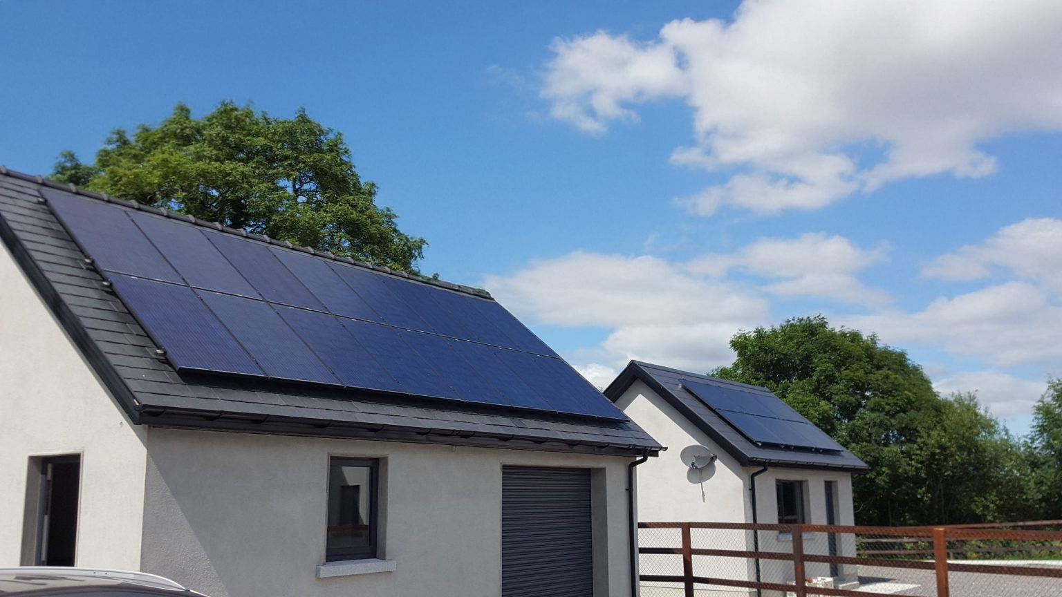 Houses with solar panels on dark gray roofs against a blue sky with fluffy clouds.