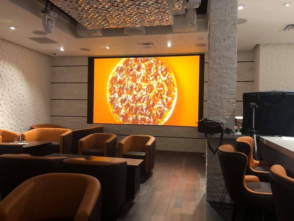 Restaurant interior with large screen displaying a pizza. Orange seating surrounds dark tables. 