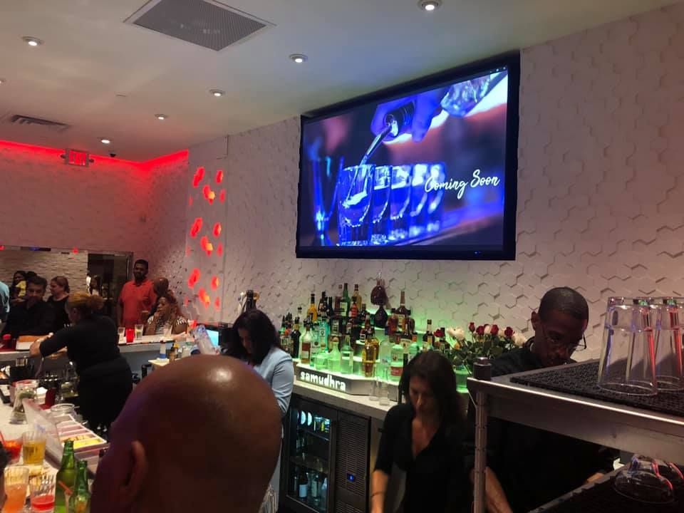 Bar interior with patrons and bartenders. TV screen shows drink pouring. Red and white décor with lit bottles.