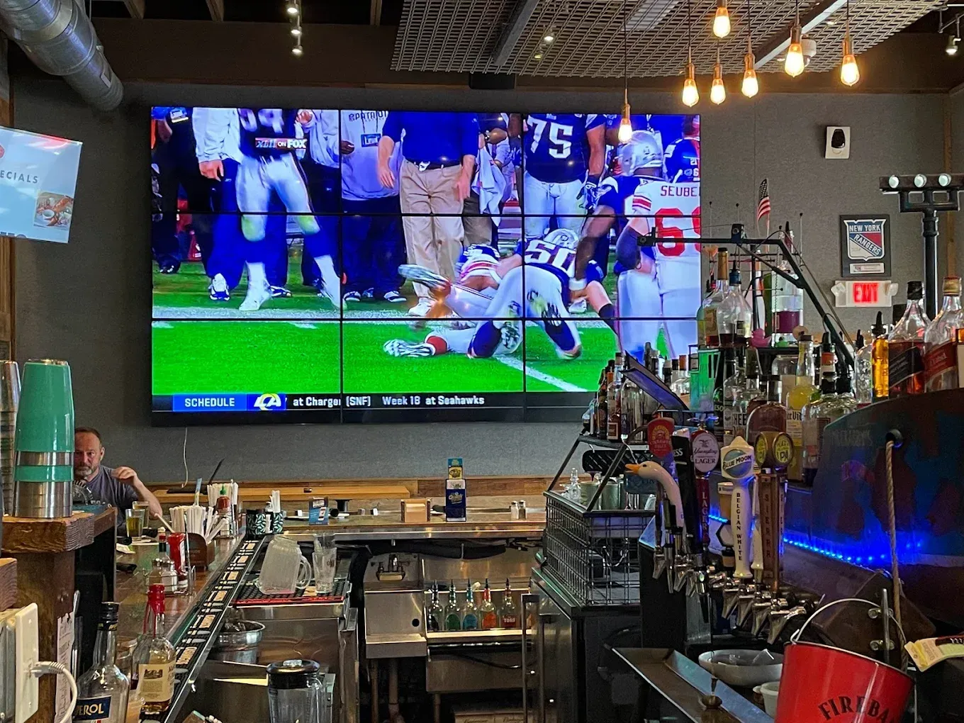 Bar interior with a large screen displaying a football game; bartender serving drinks.