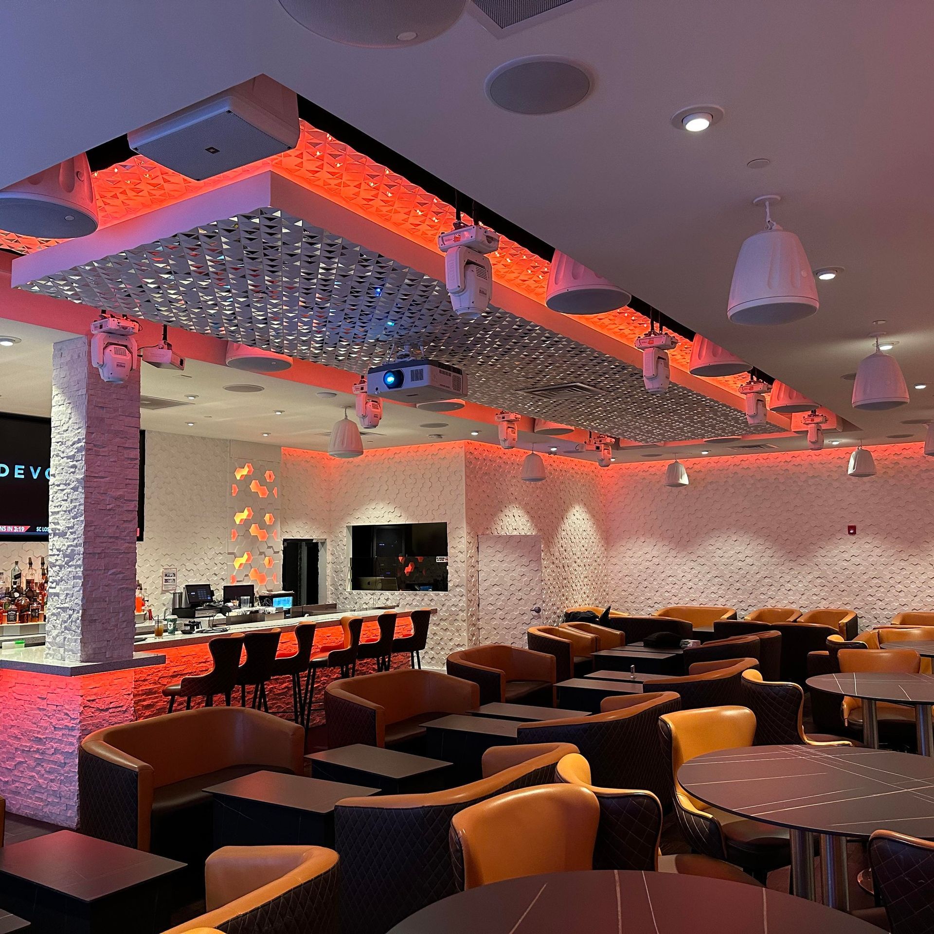 Interior of a modern bar with orange and white decor, tables, chairs, and overhead lighting.