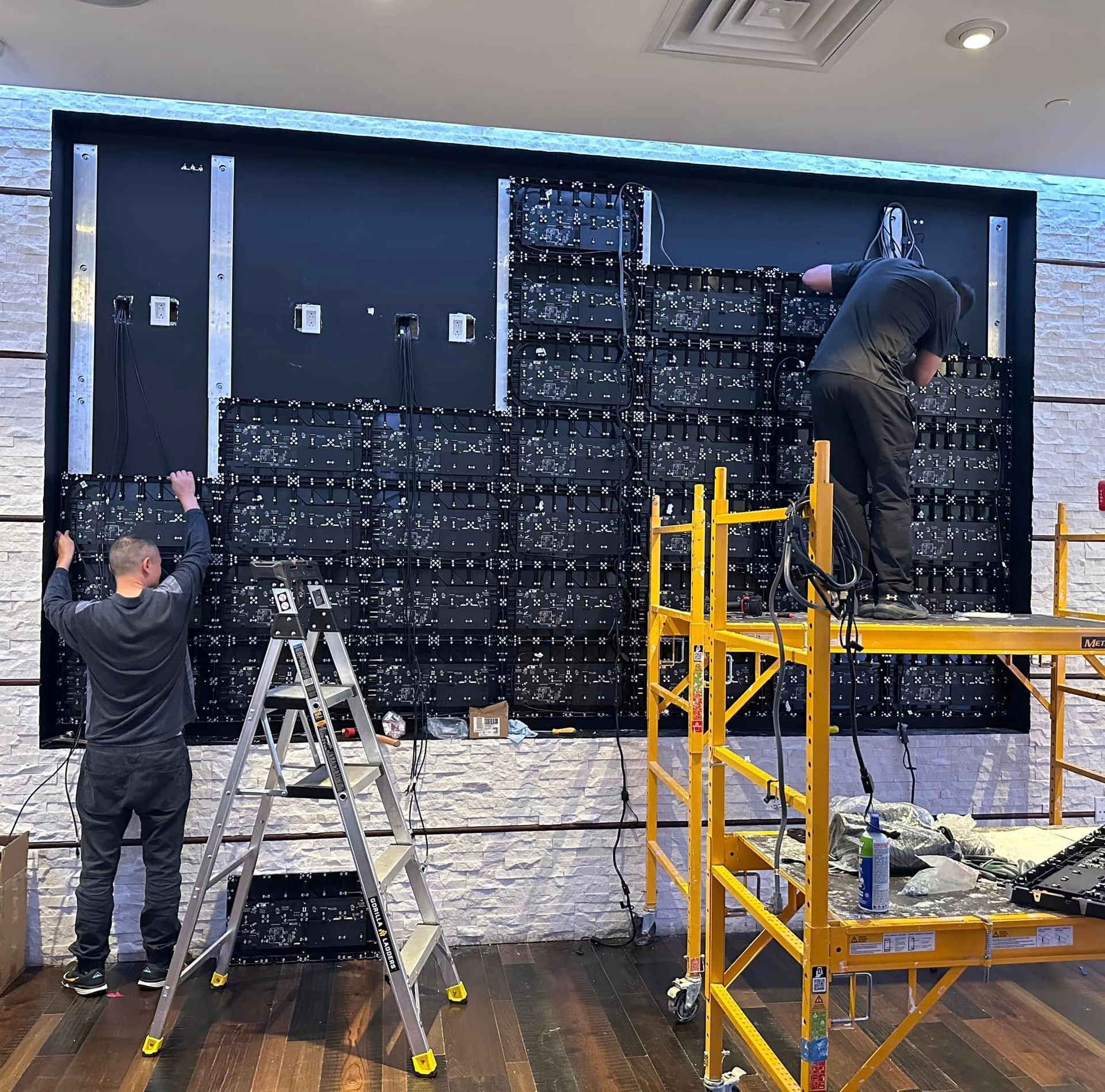 Two workers install LED display panels on a wall, using a ladder and scaffolding.