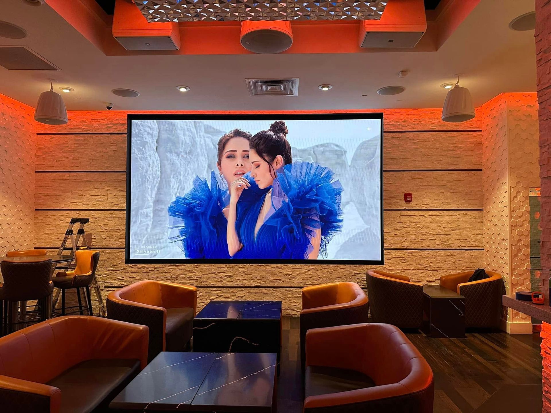 Interior lounge with large screen showing two women in blue. Orange lighting, seating.