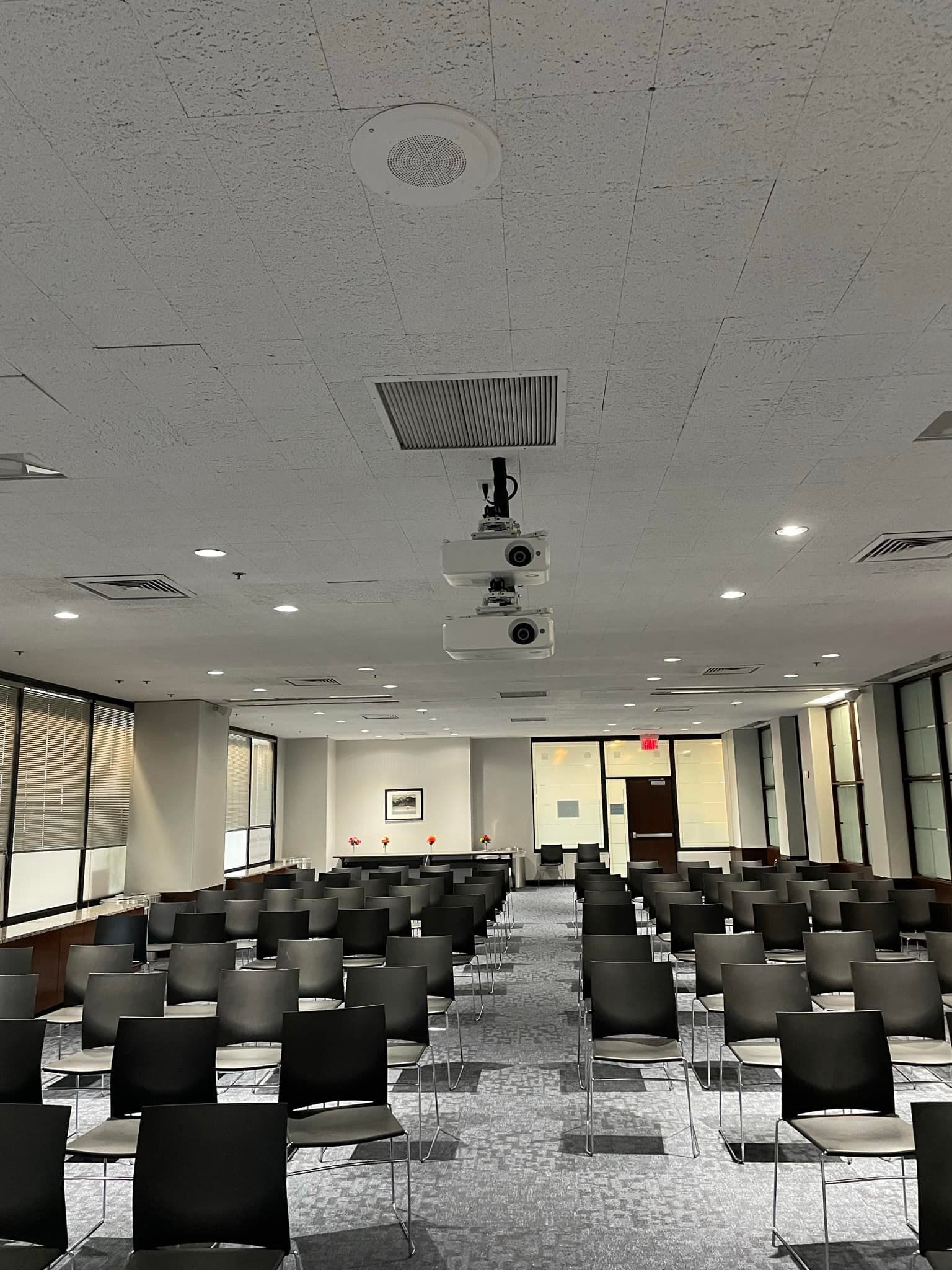 Conference room with rows of chairs, projectors hanging from the ceiling, and windows on the sides.