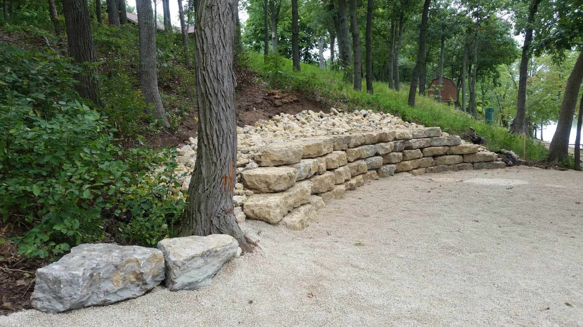 A stone wall is surrounded by trees and gravel in the middle of a forest.