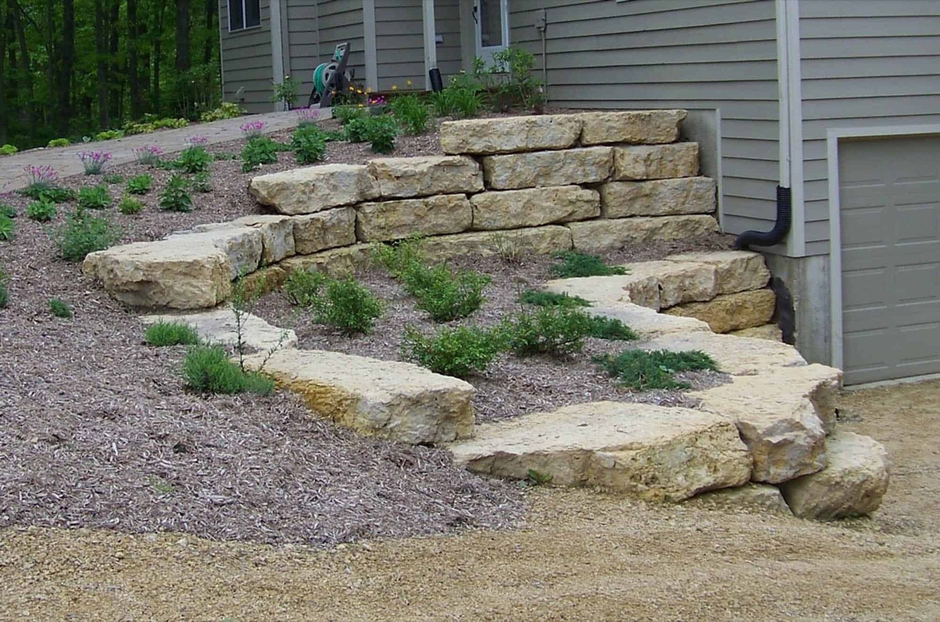 A stone wall is surrounded by gravel and plants in front of a house.