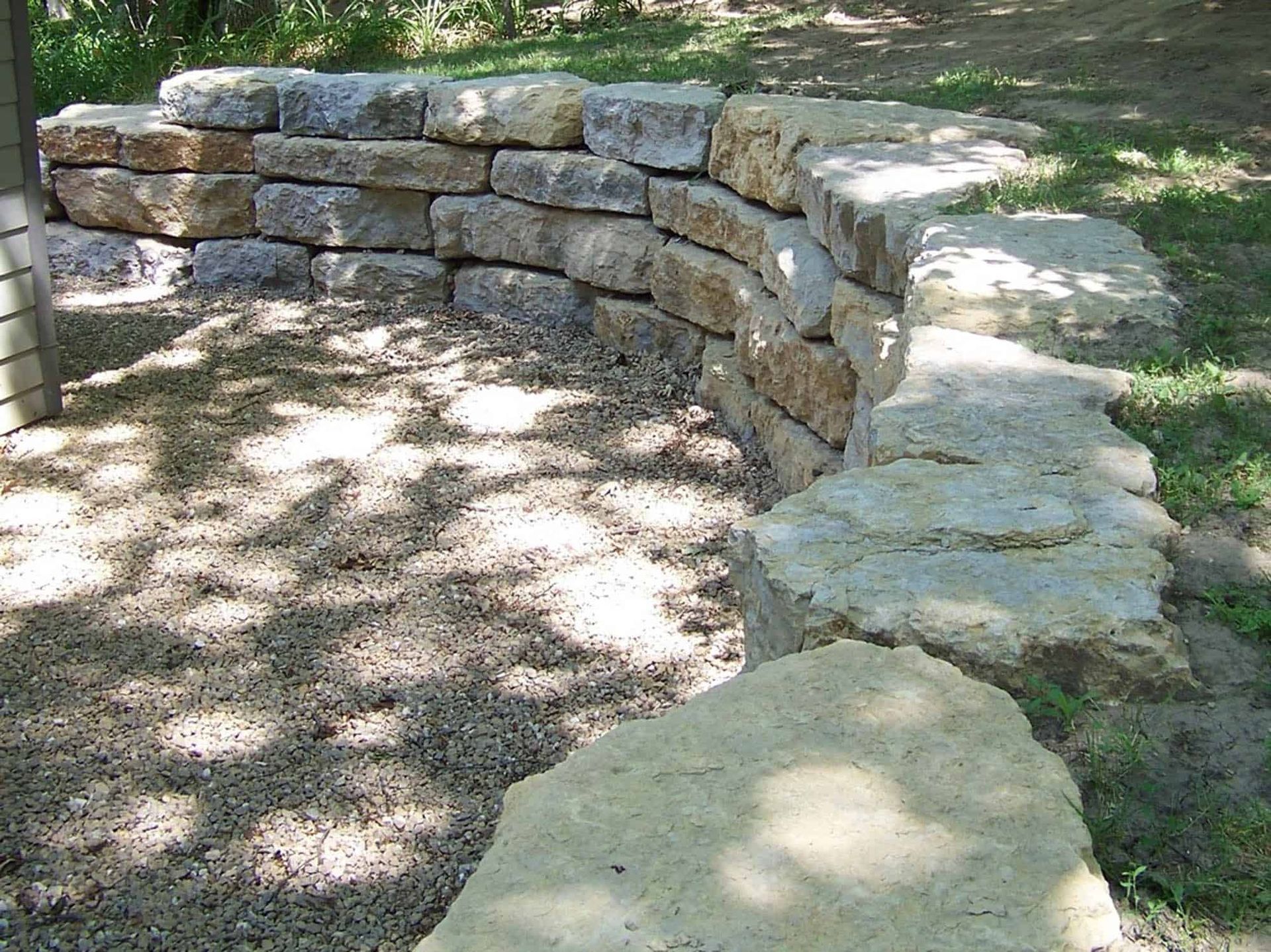 A stone wall is surrounded by gravel and grass in a yard.