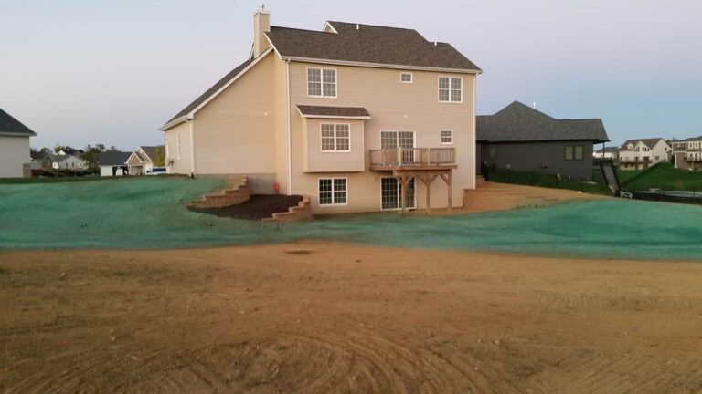A large white house with a deck is surrounded by grass and dirt.