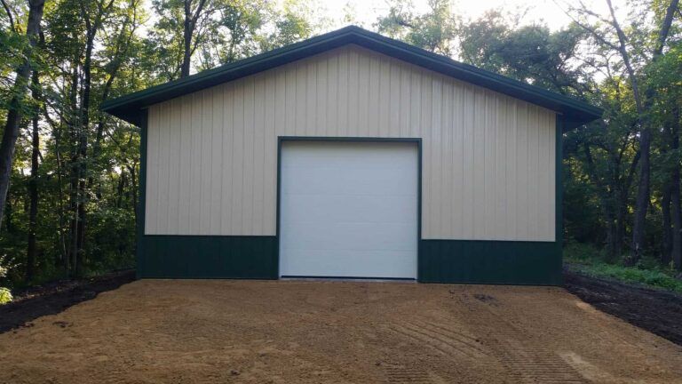 A white and green metal building with a garage door in the middle of a forest.