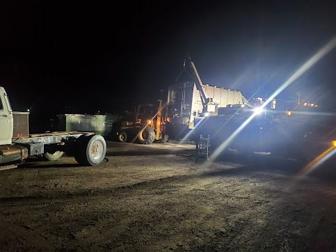 A semi-truck and heavy machinery at night. A loader is maneuvering a large object, possibly a tank, onto the truck.