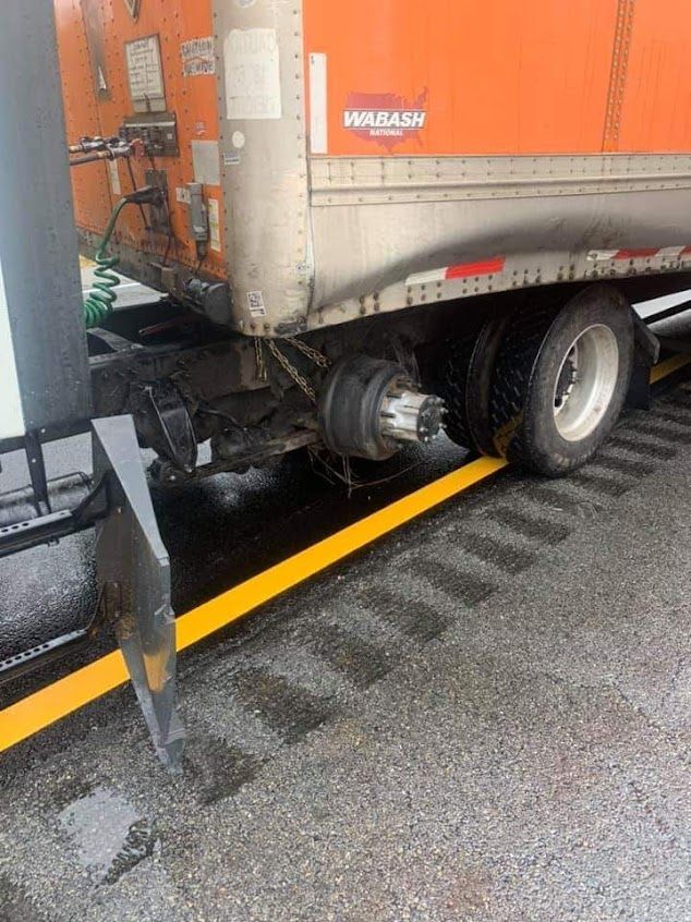 Rear section of an orange and silver semi-truck trailer with a missing tire, parked on a wet, gray surface.