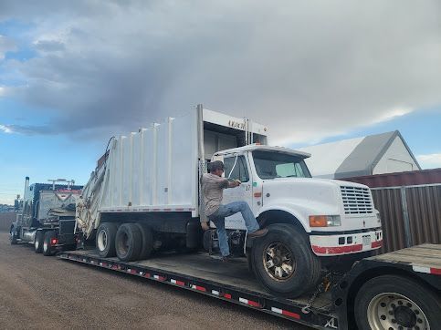 A man climbs into the cab of a white garbage truck loaded on a flatbed trailer. A second truck is parked nearby. Cloudy sky.