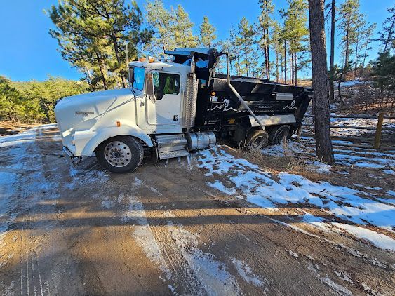 White dump truck stuck on a snow-covered dirt road in a wooded area under a blue sky.