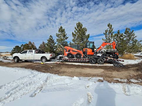 White pickup truck towing an excavator on a trailer through a snowy landscape under a blue sky.