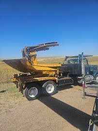 A yellow tree spade mounted on a truck in a field under a blue sky. The spade is raised in the air.