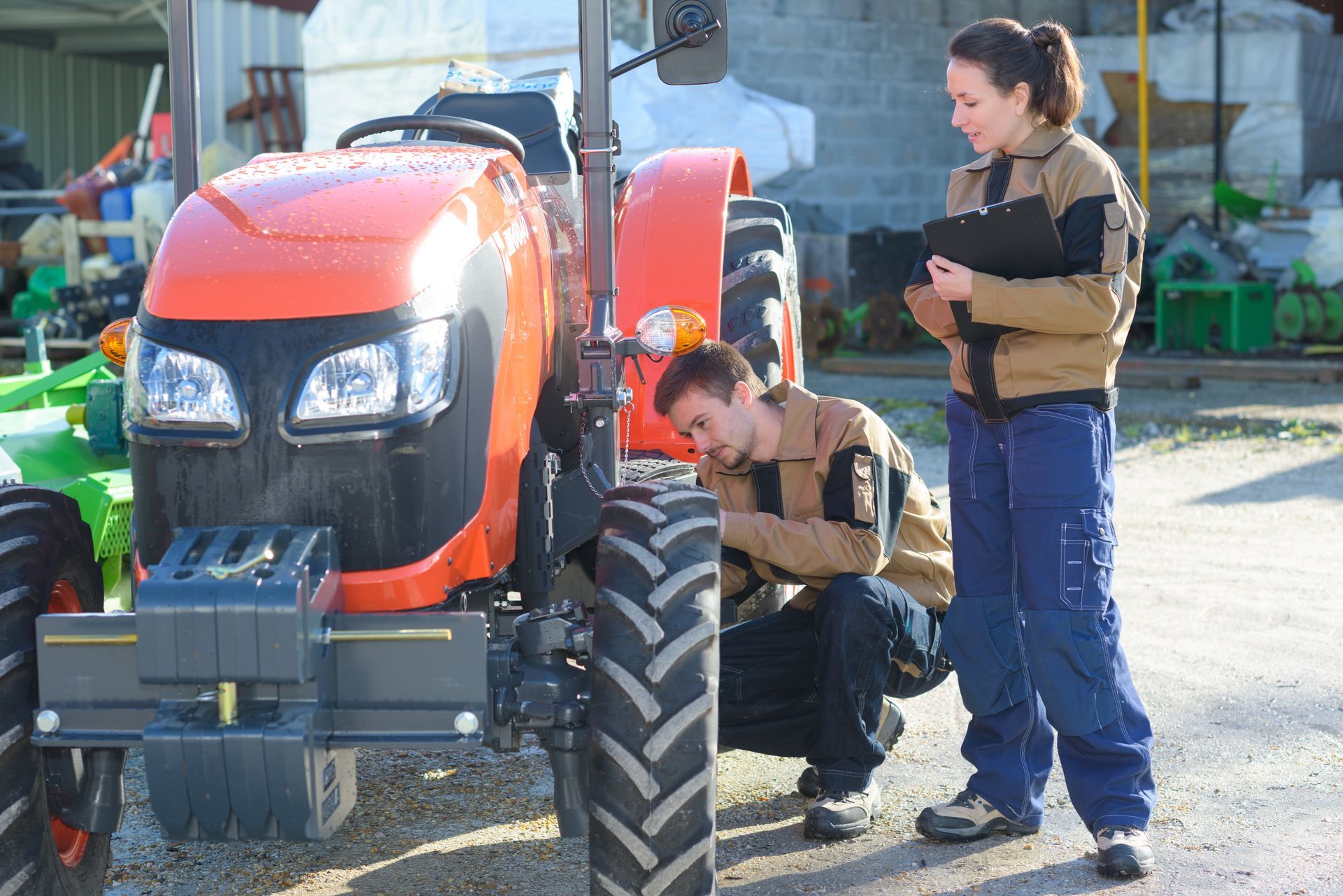 Two people examining an orange tractor outdoors. One kneels by a tire, while the other holds a clipboard.