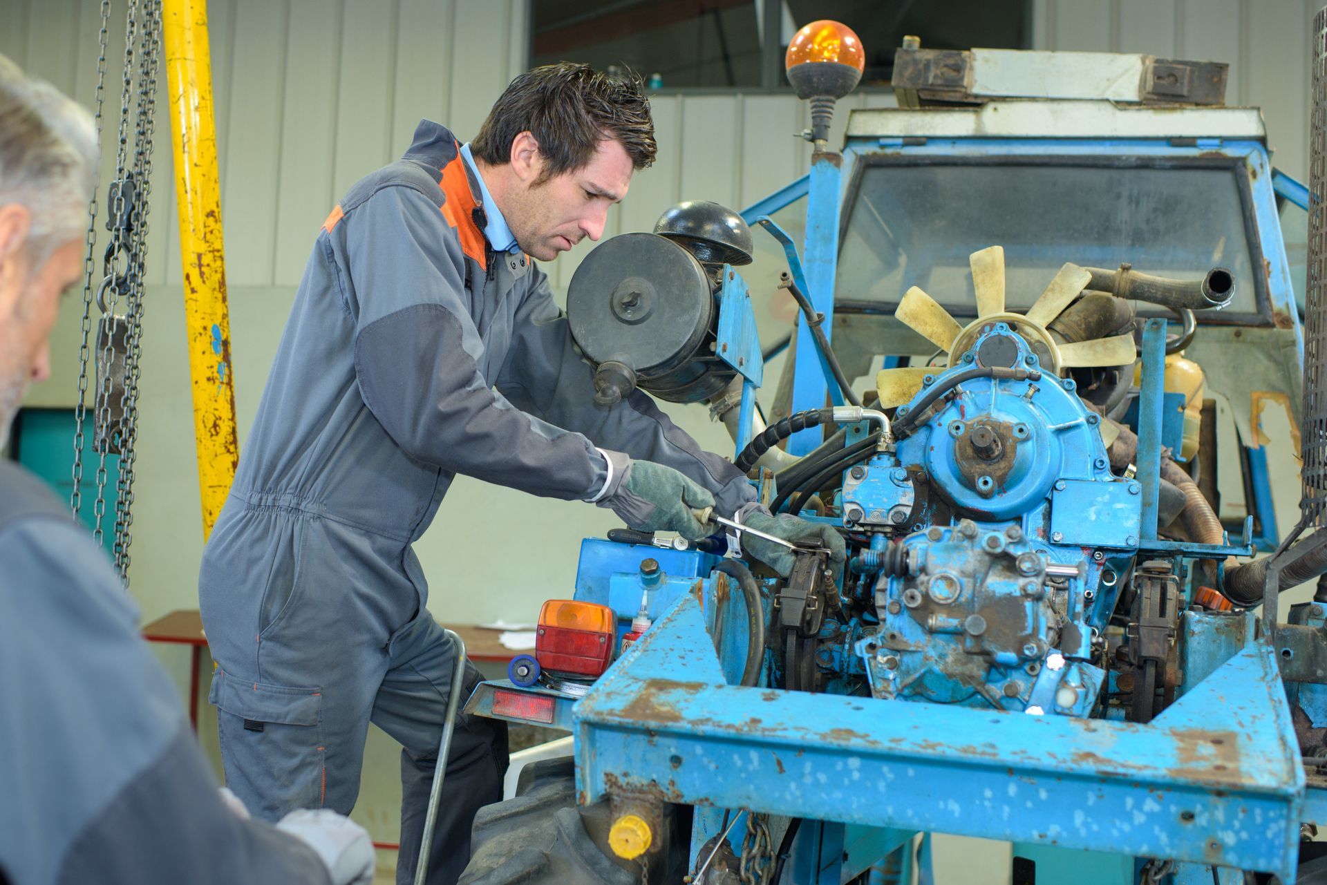 Mechanic in gray coveralls works on a blue tractor engine in a garage, while another man watches from the side.