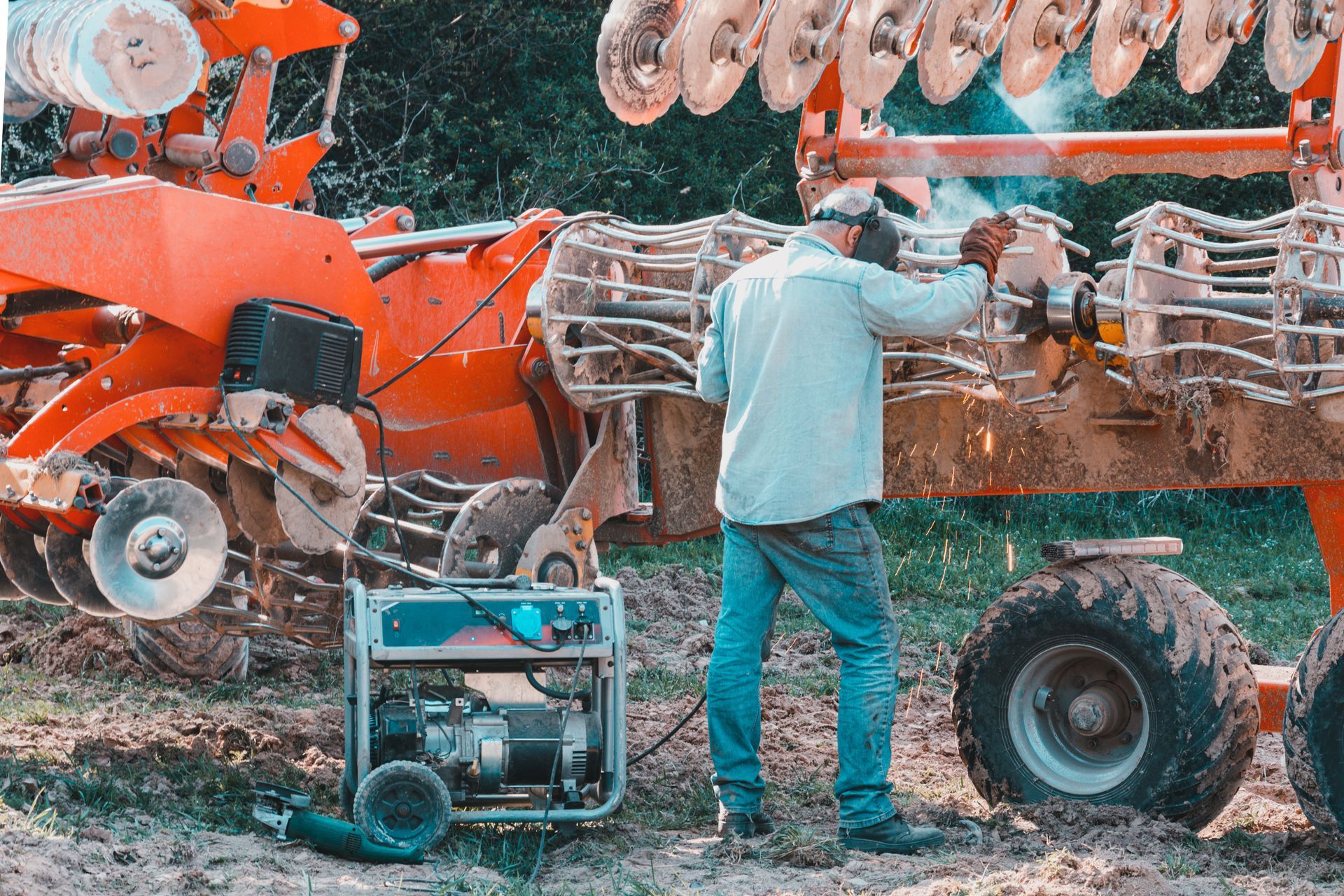 A person welds a large piece of farm machinery outdoors, near a running generator. Sparks fly.