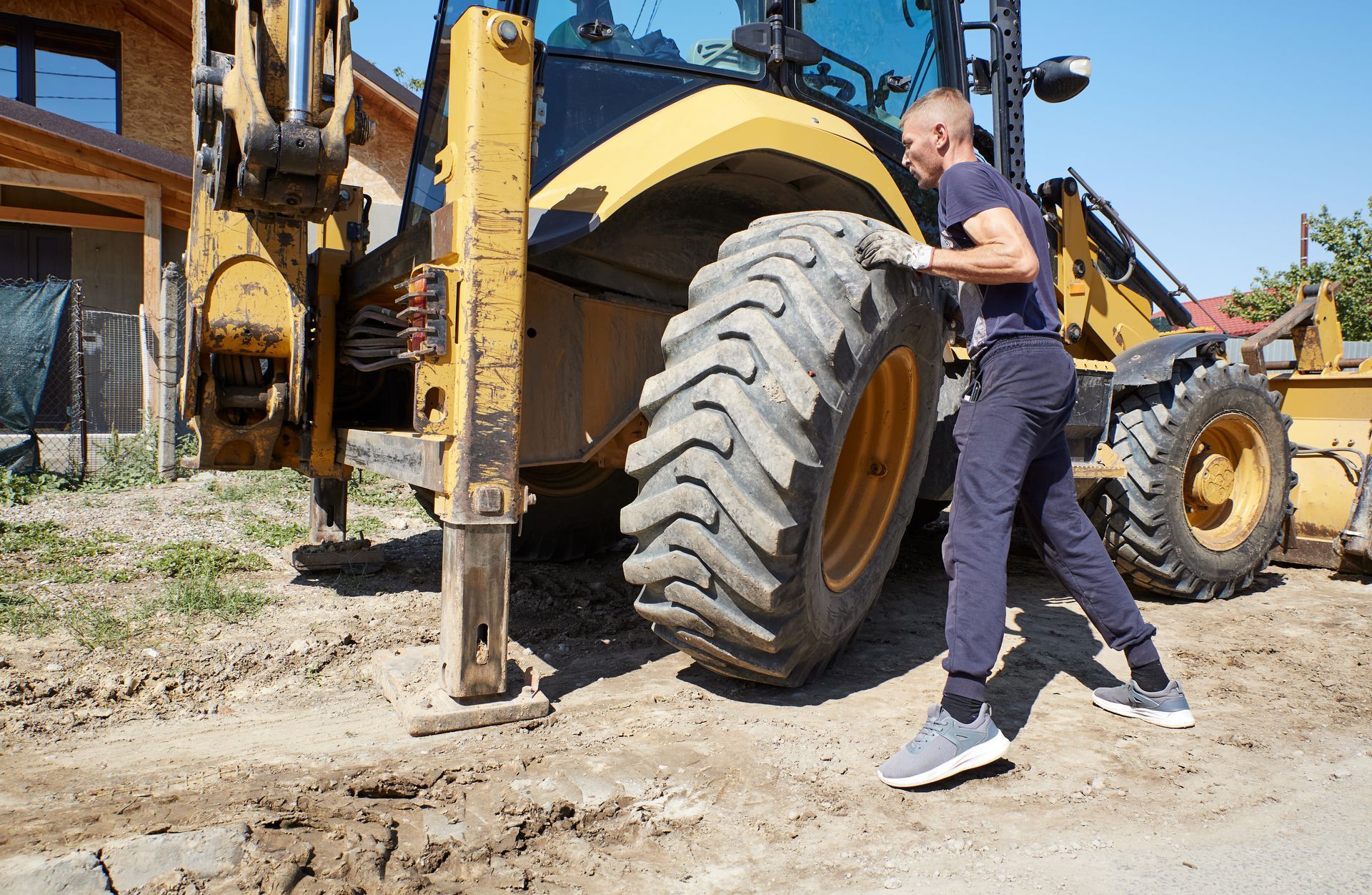 A man is changing a large, yellow construction vehicle's tire outdoors. He's lifting the tire with effort on a dirt surface.