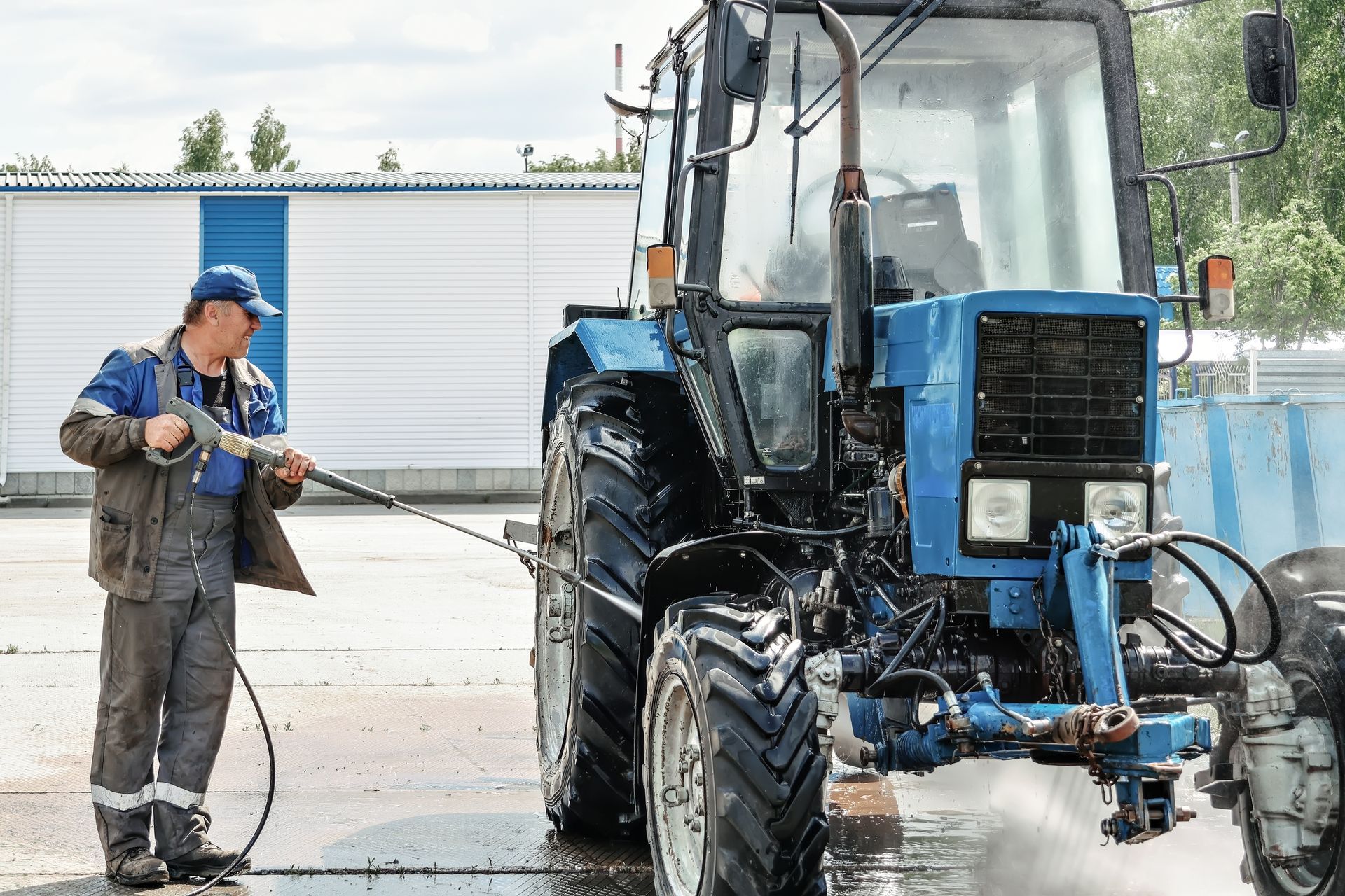 Man power washes a blue tractor in an outdoor setting. He wears work clothes and holds a pressure washer.