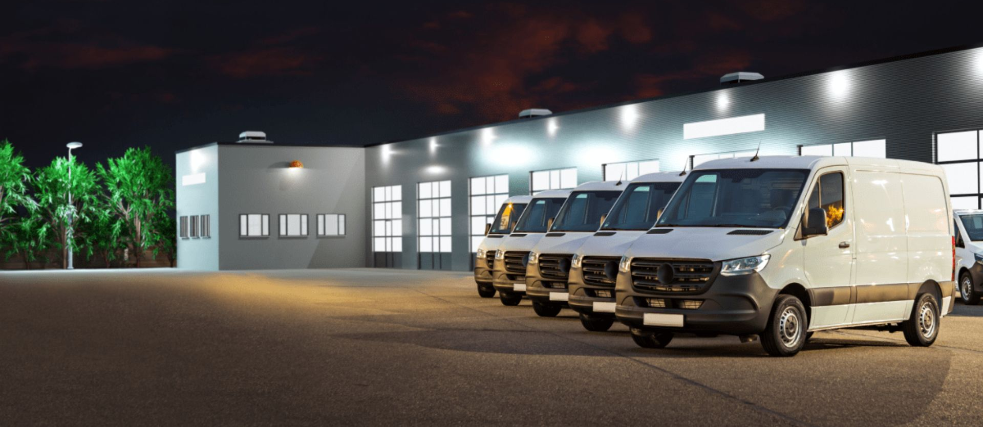 Several white vans parked outside a lit industrial building at night | Adaptive Fleet Management