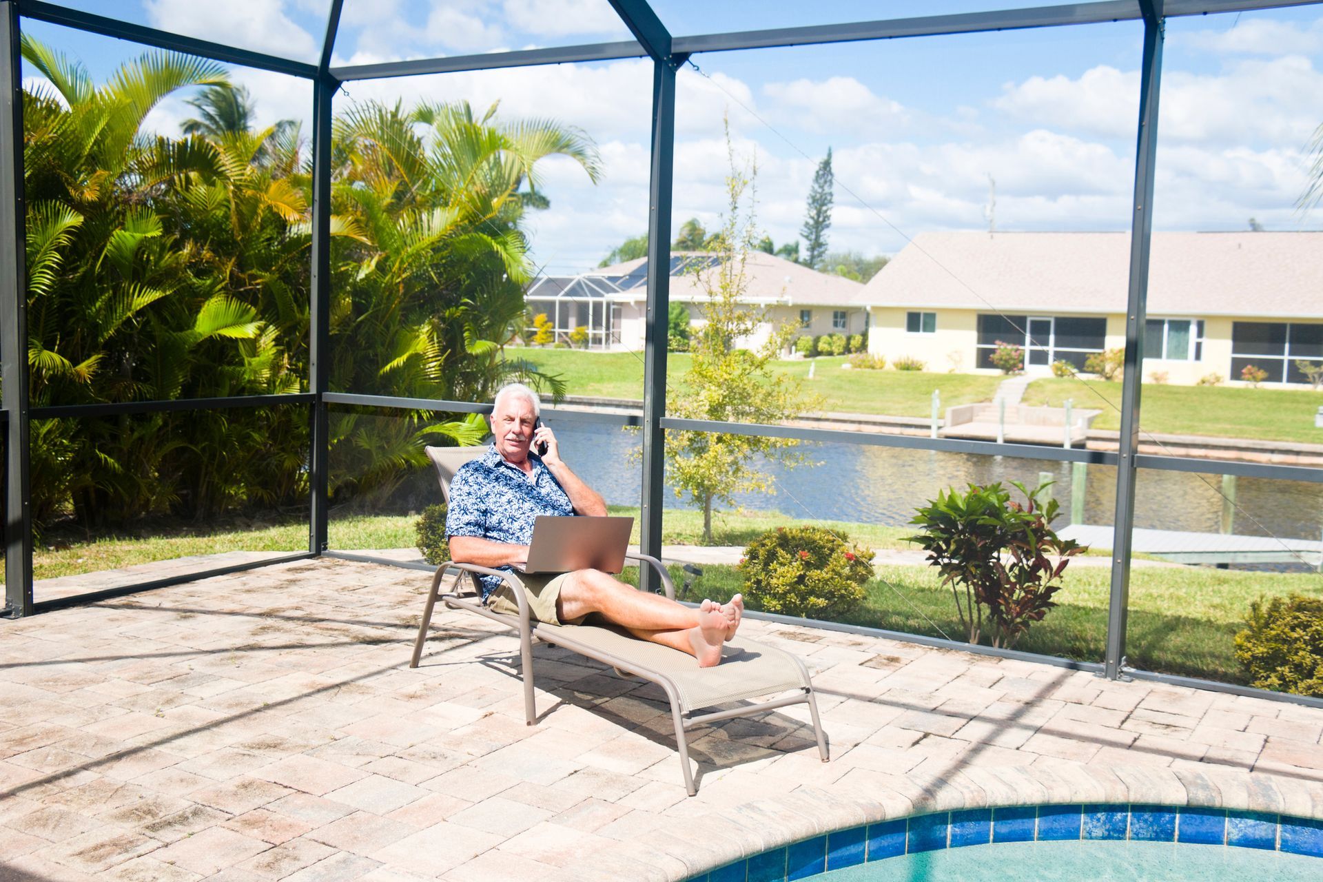 A man is sitting on a lounge chair using a laptop and talking on a cell phone