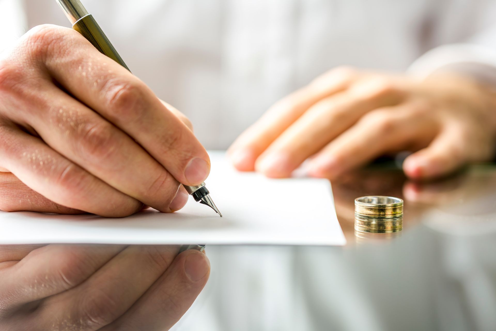 A person signs a document next to a removed wedding ring on a reflective surface.