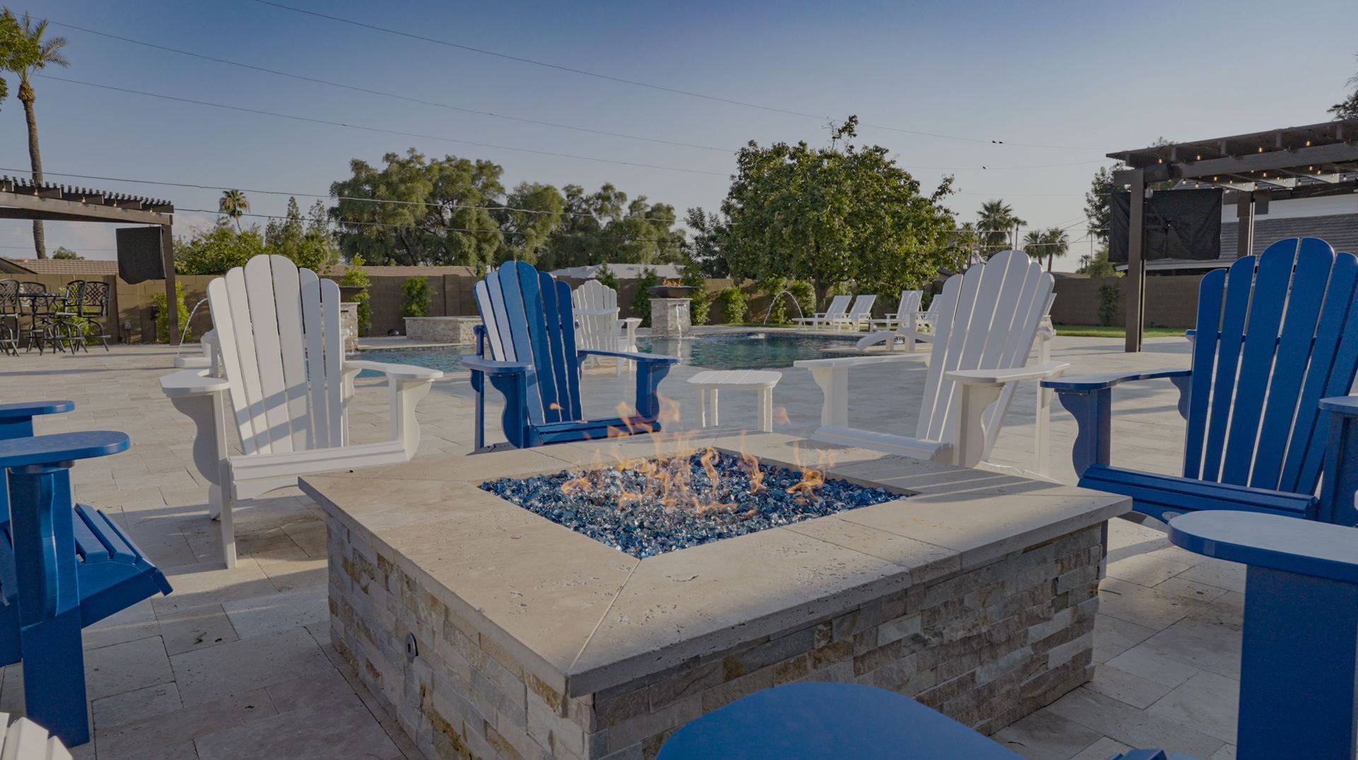 A fire pit surrounded by blue and white chairs next to a pool.