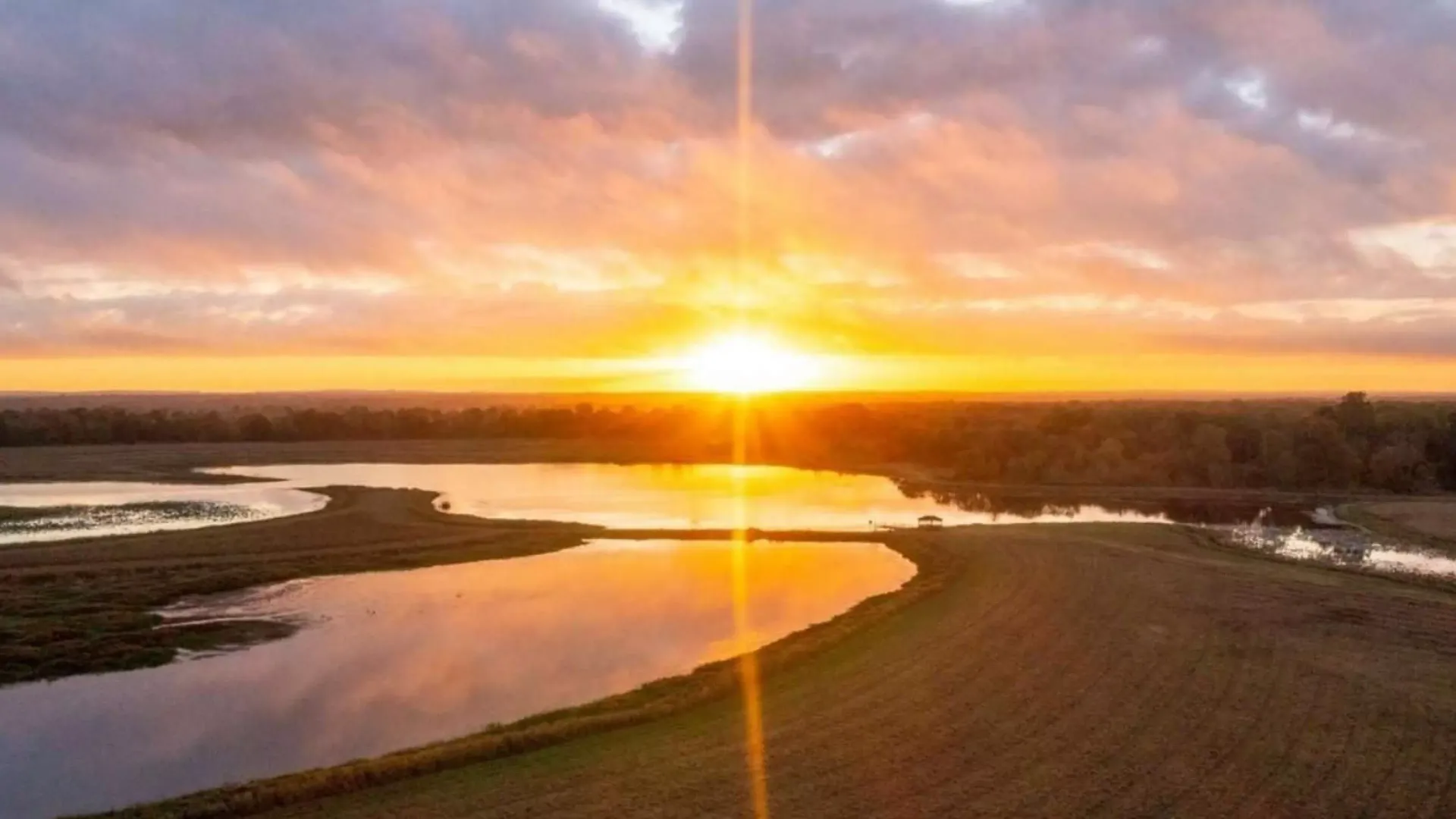 Sunset over a calm lake, reflecting golden light. Orange sky, fields, and trees in the distance.
