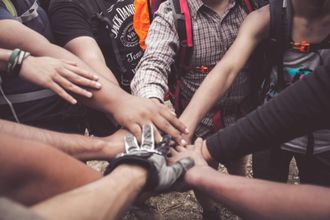 Hands stacked in a team huddle, wearing outdoor gear, conveying collaboration and unity.