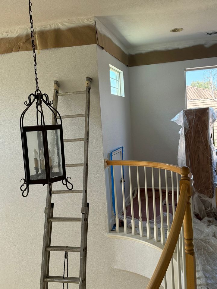 A metal ladder stands against a textured wall next to a hanging pendant lantern and a wooden staircase railing.