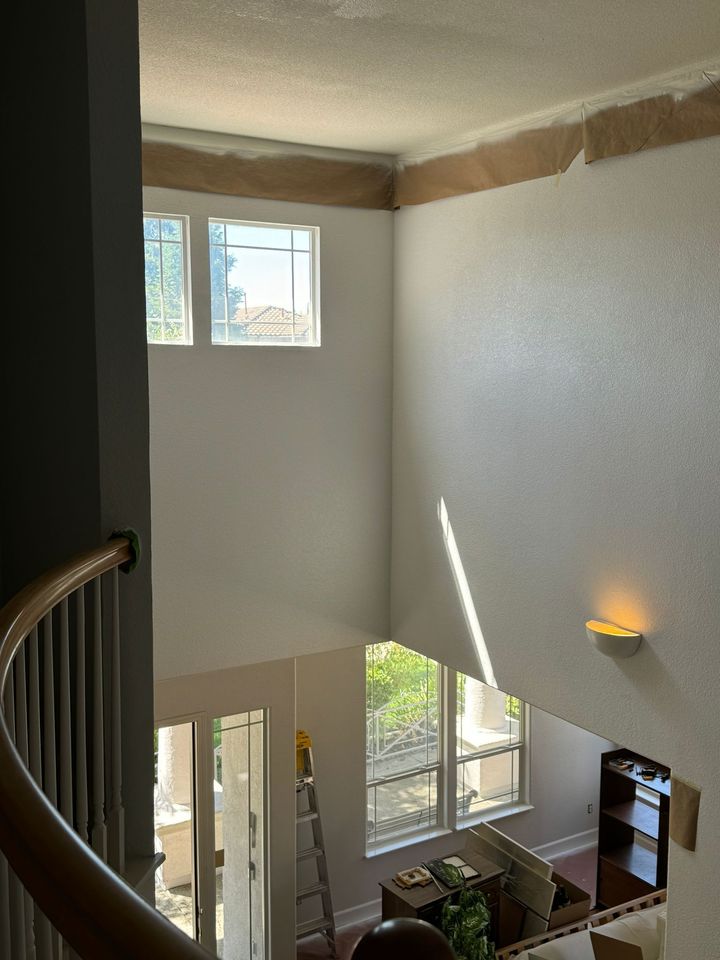 A high-angle view looking down into a living area with white walls, a vaulted ceiling, and windows in progress of painting.