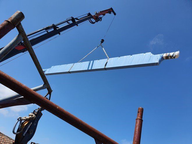 Un lungo pannello rivestito di blu, sospeso da una gru contro un cielo azzurro brillante.