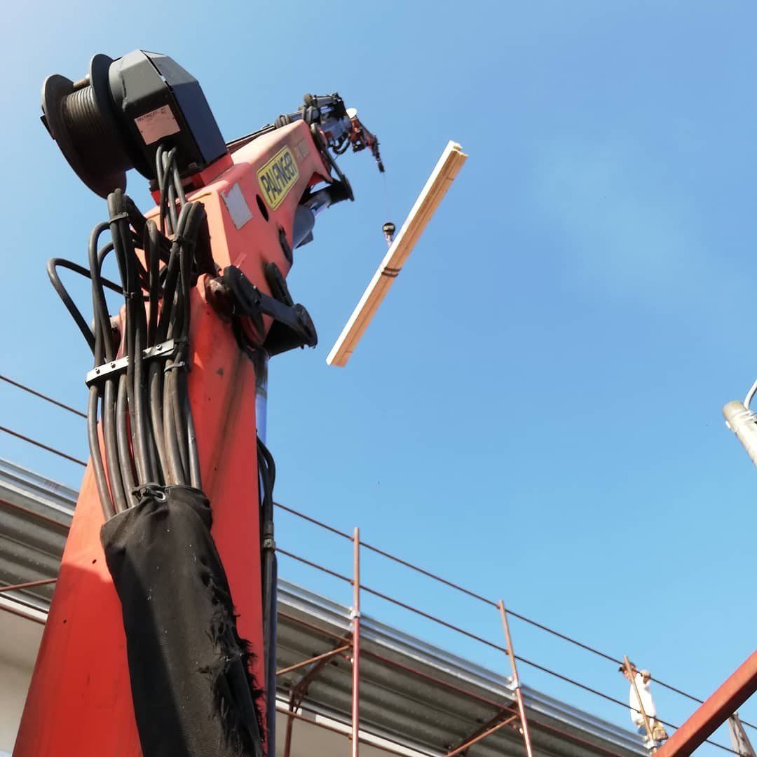 Una gru arancione solleva una trave di legno contro un cielo azzurro, vicino a un edificio con impalcature.