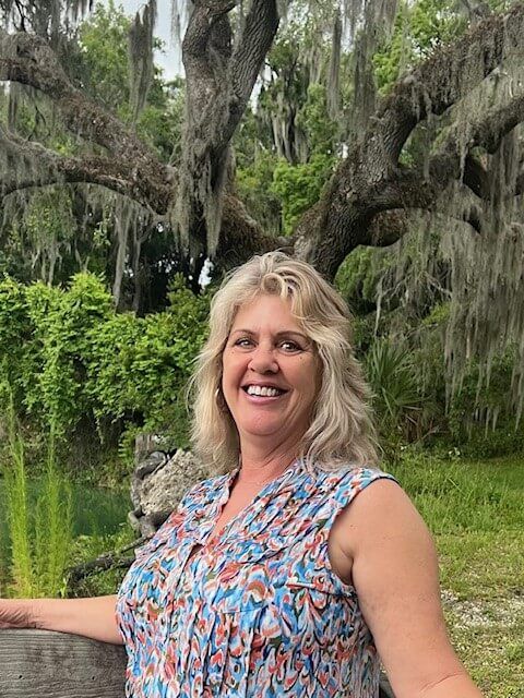 Woman with blonde hair smiles in front of a tree draped with Spanish moss.