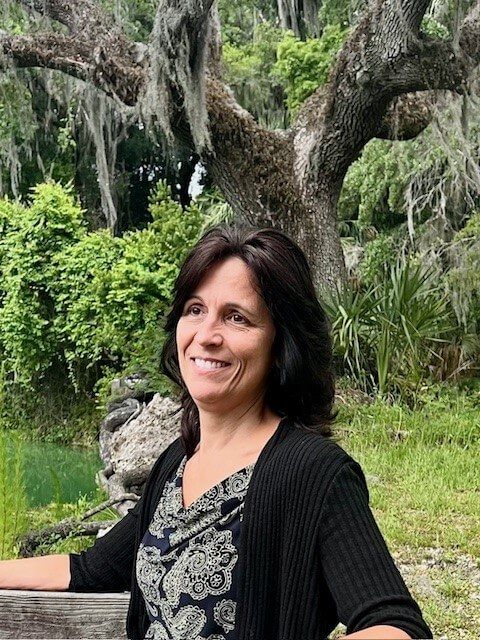 Woman with dark hair smiles outdoors, in front of a mossy tree and green foliage.