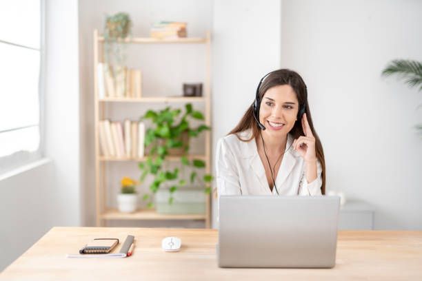 Woman with headset working on laptop at a desk, smiling; home office with plants.