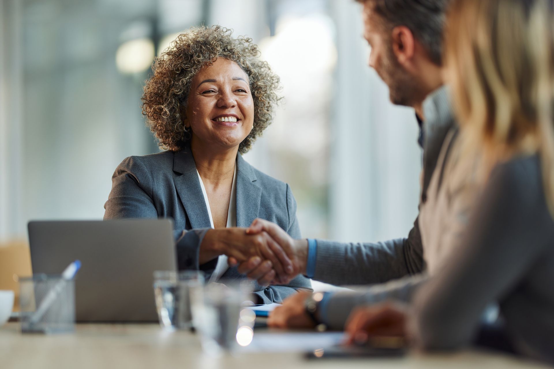 Woman shaking hands with a man, smiling, at a table with a laptop and paperwork.