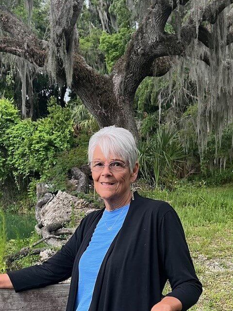 Woman with white hair, in black cardigan and blue shirt, smiles outdoors near a tree with Spanish moss.