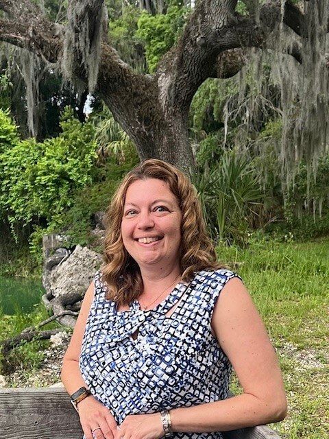 Woman smiles outdoors, wearing patterned top, under a moss-draped tree.