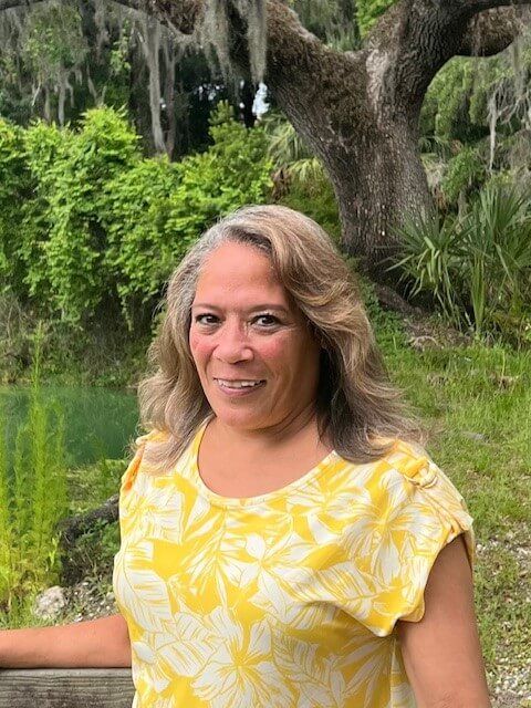Woman in yellow floral shirt smiling outside by greenery.